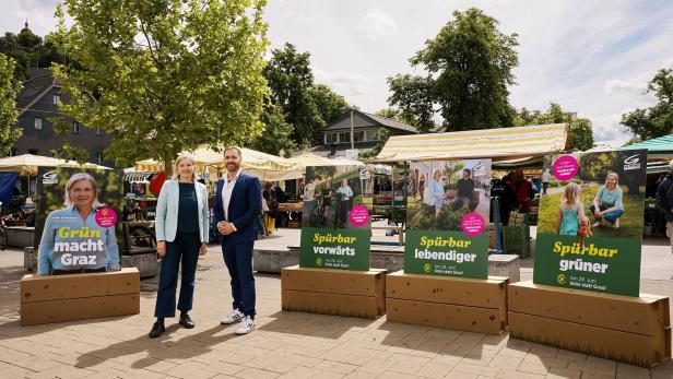 Zwei Personen stehen vor Wahlplakaten der Grünen auf einem belebten Platz.