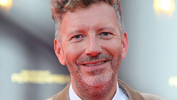 Thorsten Schorn trägt einen beigefarbenen Anzug mit Fliege und Mikrofon auf einem roten Teppich.
