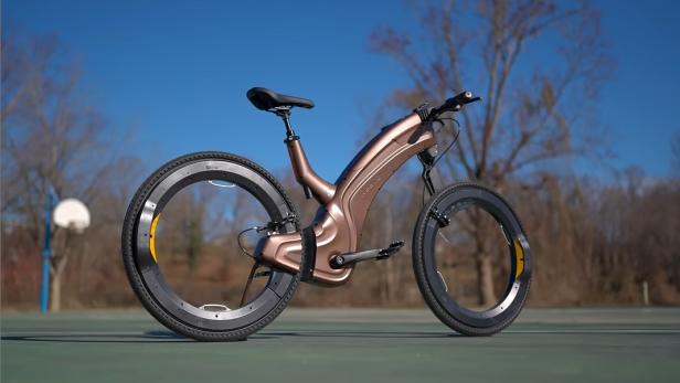 Futuristisches Fahrrad mit bronzefarbenem Rahmen und auffälligen, speichenlosen Rädern auf einem Sportplatz.