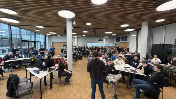 Viele Menschen sitzen und stehen in einem modernen Seminarraum mit Computern und diskutieren.