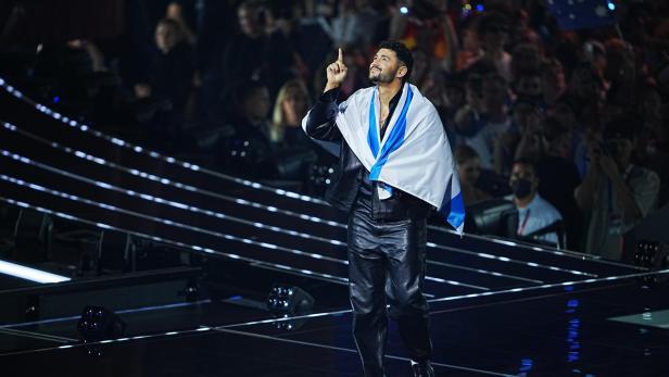 Ein Mann mit einer blau-weißen Flagge um die Schultern steht auf einer Bühne und zeigt mit dem Finger nach oben.
