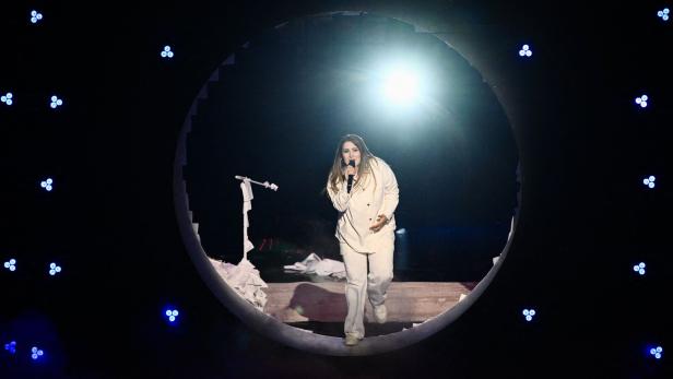 Eine Frau in weißer Kleidung singt auf einer Bühne mit kreisförmigem Durchgang und starkem Licht im Hintergrund.