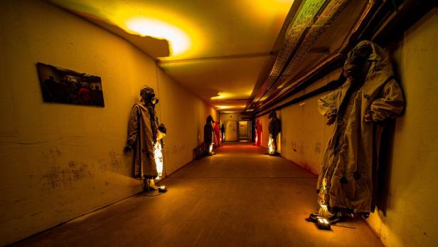 Ein langer, gelb beleuchteter Gang mit Schaufensterpuppen in Schutzanzügen und Gasmasken an den Wänden.
