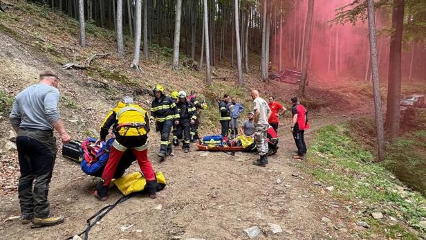 Mehrere Rettungskräfte und Helfer versorgen eine Person auf einer Trage im Wald, während im Hintergrund roter Rauch aufsteigt.