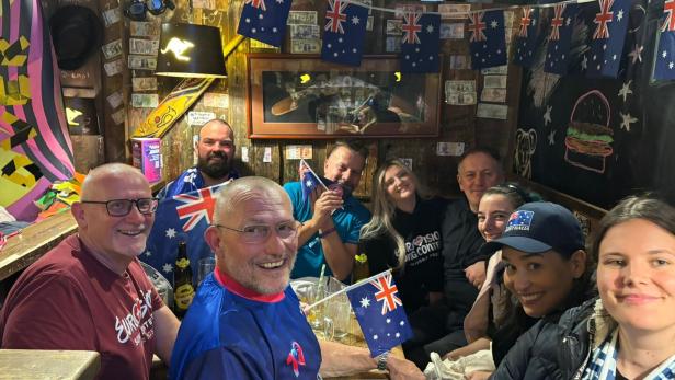 Eine fröhliche Gruppe von Menschen sitzt in einem dekorierten Raum mit vielen australischen Flaggen und lächelt in die Kamera.