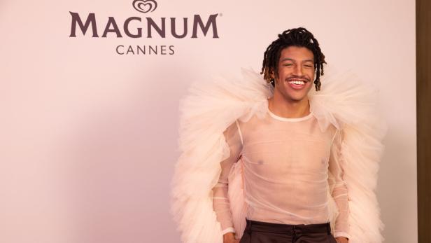 Ein lächelnder Mann mit Dreadlocks trägt ein transparentes Oberteil und einen bauschigen Kragen vor einem Magnum Cannes Logo.
