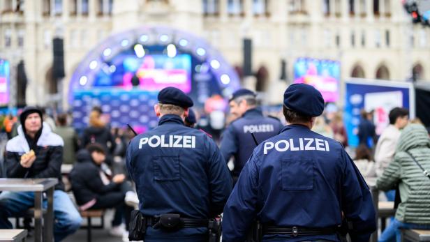 Mehrere Polizisten in Uniform überwachen eine Veranstaltung im Freien mit vielen Menschen und einer beleuchteten Bühne.