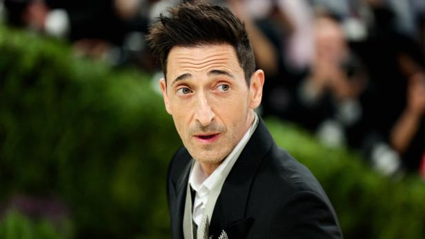Adrien Brody im schwarzen Anzug mit Brosche blickt aufmerksam zur Seite.