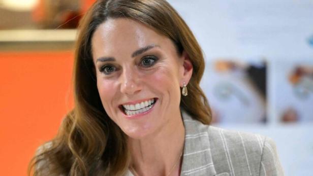 Kate, mit Blazer, lächelt und gestikuliert mit beiden Händen.