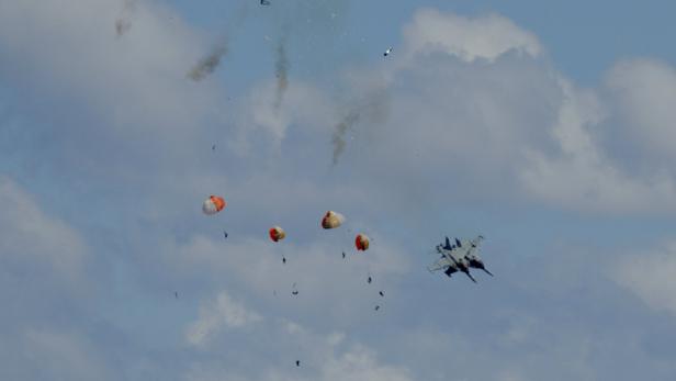 Ein Kampfjet fliegt neben mehreren fallenden Objekten mit Fallschirmen und Rauchspuren am Himmel.