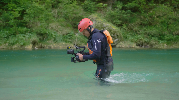 Eine Person mit Helm und Kamera steht im flachen Wasser eines türkisfarbenen Flusses