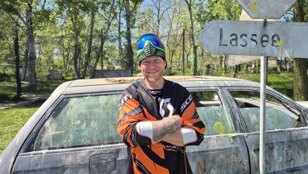 Mann in Paintball-Ausrüstung steht vor einem alten Auto und einem Wegweiser nach Lassee.