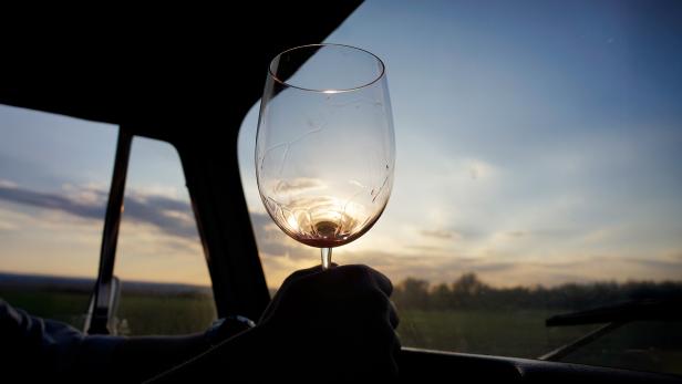 Eine Person hält bei Sonnenuntergang ein leeres Weinglas vor ein Fahrzeugfenster.