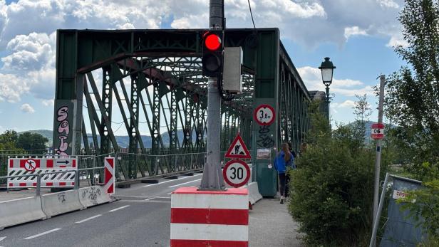 Eine grüne Stahlbrücke mit Baustellenabsperrungen, Verkehrsschildern und roter Ampel, zwei Personen gehen auf dem Gehweg.