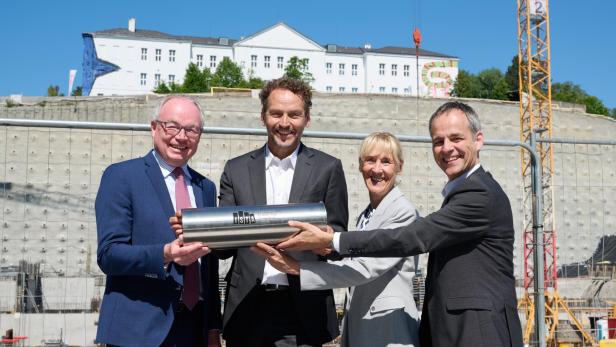 Vier Personen im Anzug halten gemeinsam eine silberne Röhre vor einer Baustelle und lächeln in die Kamera.