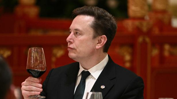 Ein Mann im Anzug hält ein Glas Rotwein und blickt nach links, im Hintergrund rote Dekoration.