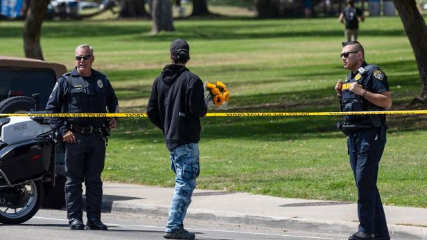Zwei Polizisten sprechen mit einer Person, die einen Strauß Sonnenblumen hält, hinter einer Absperrung im Park.