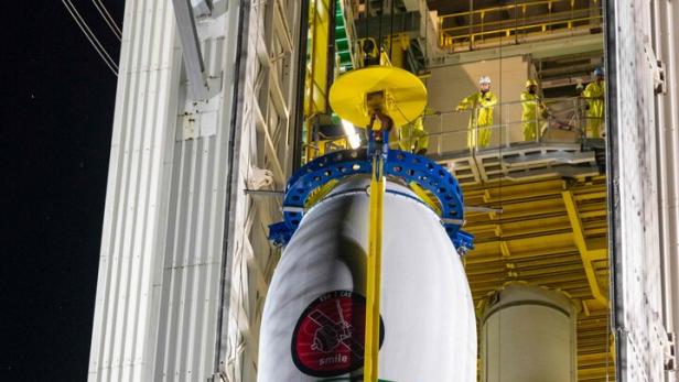 Eine Raketenstufe wird in einer Montagehalle mit gelben Gerüsten und Technikern in Schutzanzügen vorbereitet.