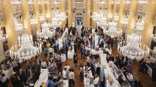 Viele Menschen besuchen eine Messe mit Ständen in einem prunkvollen Saal mit großen Kronleuchtern und goldenen Säulen.