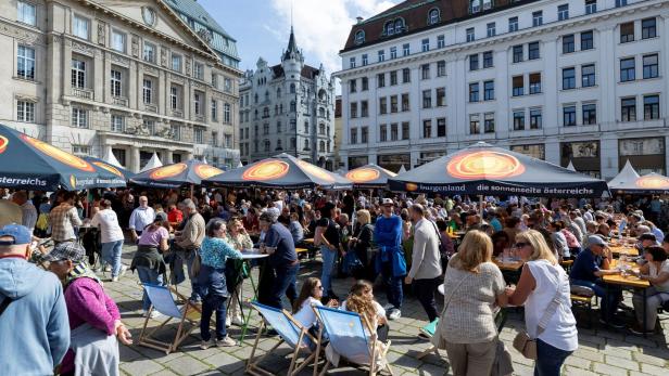 Viele Menschen sitzen und stehen an Tischen unter Sonnenschirmen auf einem belebten Platz inmitten historischer Gebäude.