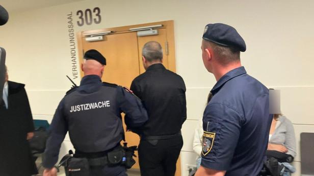 Zwei Justizwachebeamte begleiten einen Mann in schwarzer Kleidung zu einem Gerichtssaal mit der Nummer 303.