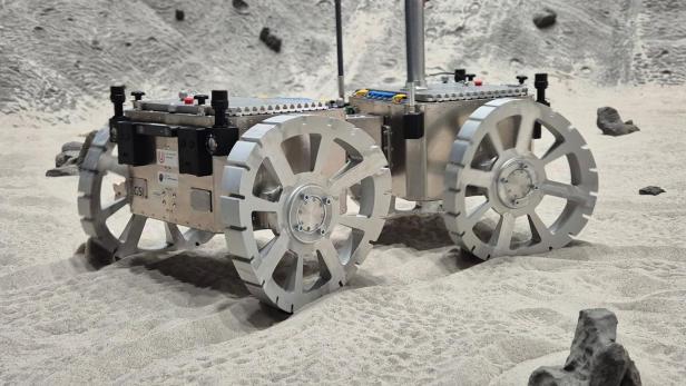 Mars-Rover steht auf sandigem Untergrund des Testgeländes. Seine silbernen Räder sind gebogen.