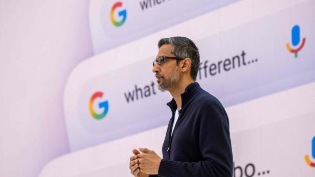 Ein Mann spricht vor einer großen Leinwand mit Google-Suchanfragen und buntem Mikrofon-Symbol.