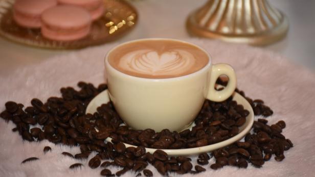 Cappuccino mit Latte Art in einer Tasse, umgeben von Kaffeebohnen und rosa Macarons im Hintergrund.