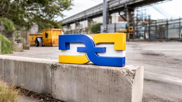 Blau-gelbes DDZ-Logo auf Betonmauer vor Industrie- und Baustellenkulisse.