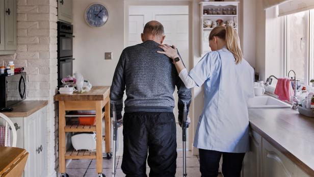 Pflegekraft unterstützt älteren Mann mit Gehhilfen in einer hellen Küche.