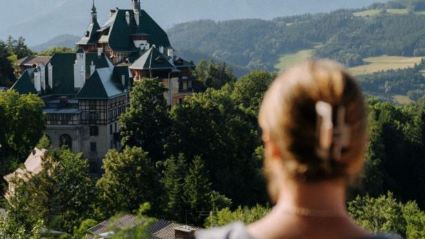 Blick über die Schulter einer Frau mit zusammengebundenem Haar auf ein Schloss in einer bewaldeten Hügellandschaft.