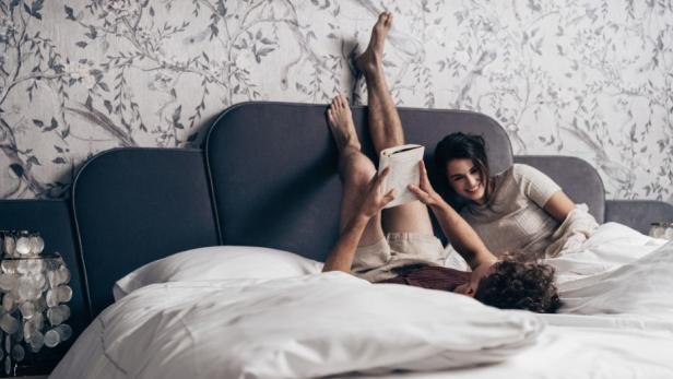 Zwei Personen liegen auf einem Bett mit weißer Bettwäsche vor einer Wand mit floralem Muster, eine Person hält ein Buch, die andere lehnt am Kopfteil.