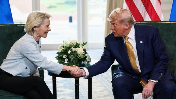 Ursula von der Leyen und Donald Trump geben sich die Hand vor Flaggen.
