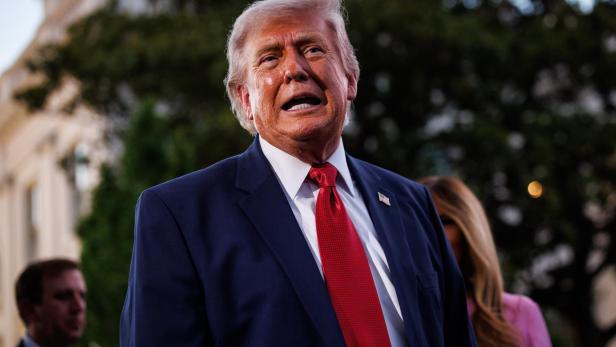 Donald Trump spricht in einem dunklen Anzug mit roter Krawatte vor einem Gebäude.