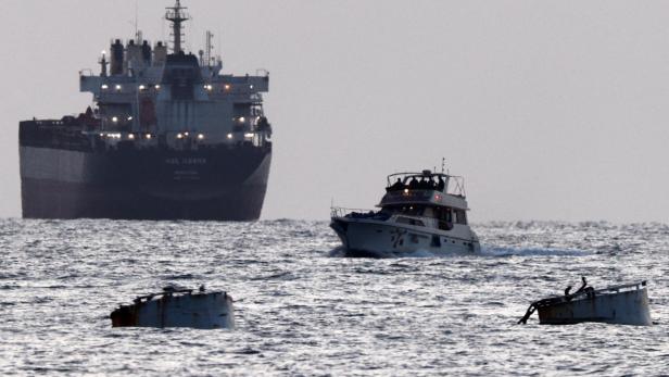 Ein Frachtschiff und ein kleineres Boot sind auf dem Meer zu sehen, im Vordergrund treiben Trümmer.