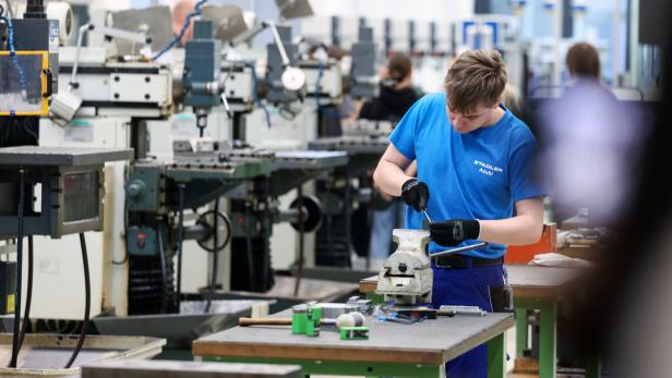 Ein junger Mann arbeitet konzentriert an einem Schraubstock in einer Werkstatt mit mehreren Maschinen im Hintergrund.