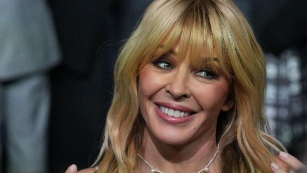 Kylie Minogue mit blondem Haar und Pony lächelt, trägt eine Perlenkette und steht vor unscharfem Hintergrund.