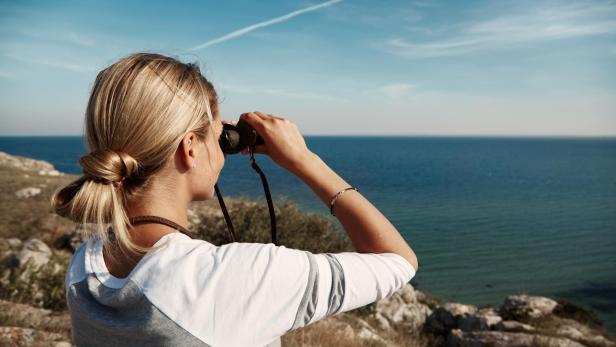 Frau mit Fernglas blickt von einer Küste aus über das Meer in die Ferne.