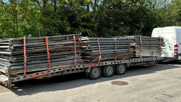 Ein weißer Transporter zieht einen Anhänger mit gestapelten Metallteilen, die mit Gurten gesichert sind.