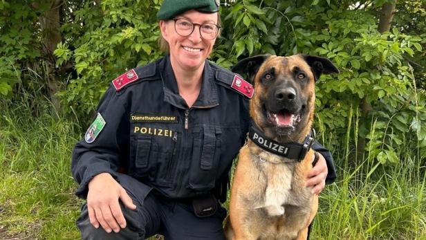 Eine Polizistin in Uniform kniet neben einem Polizeihund, beide lächeln und sitzen vor grünem Gebüsch.