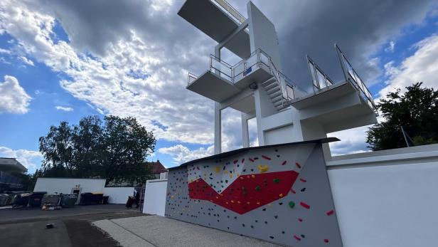 Ein hoher Sprungturm mit mehreren Plattformen steht neben einer bunten Kletterwand im Freien unter bewölktem Himmel.