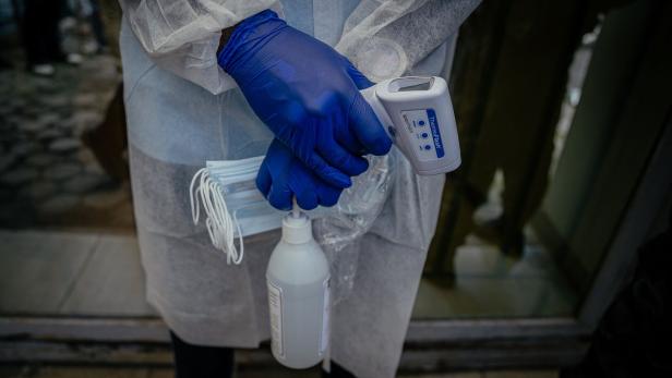 Eine Person mit blauen Handschuhen hält einen Infrarot-Thermometer, Desinfektionsmittel und mehrere OP-Masken.