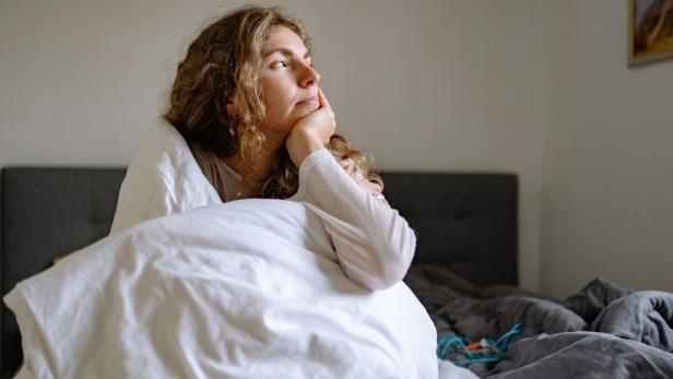 Eine Frau liegt nachdenklich unter einer weißen Decke im Bett und blickt aus dem Fenster.