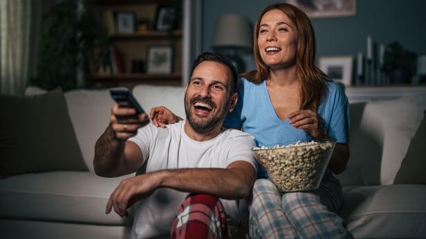 Ein lachendes Paar sitzt in Pyjamas auf dem Sofa, schaut fern und isst gemeinsam Popcorn.