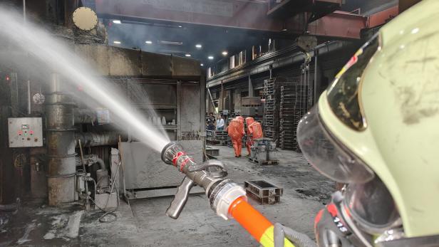Ein Feuerwehrmann spritzt Wasser in einer Industriehalle, im Hintergrund arbeiten Personen in orangefarbenen Schutzanzügen.