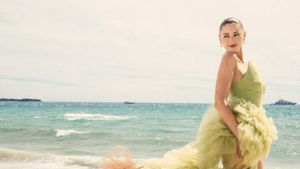 Eine Frau steht am Strand vor dem Meer und trägt ein auffälliges, voluminöses Kleid aus grünem und braunem Tüll.