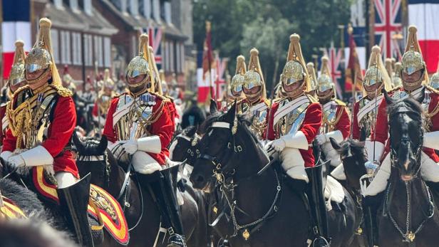 Britische Soldaten in Paradeuniform reiten auf schwarzen Pferden durch eine festlich geschmückte Straße.