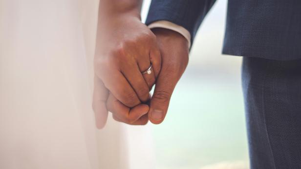 Zwei Menschen in Hochzeitskleidung halten sich an den Händen, ein Verlobungsring ist sichtbar.
