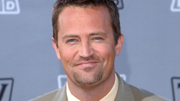 Matthew Perry lächelt mit kurzem, dunklem Haar und leichtem Bart vor grauem Hintergrund.
