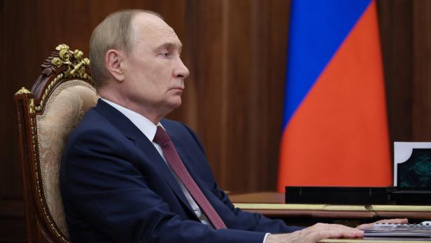Putin sitzt am Tisch, im Hintergrund die russische Flagge.